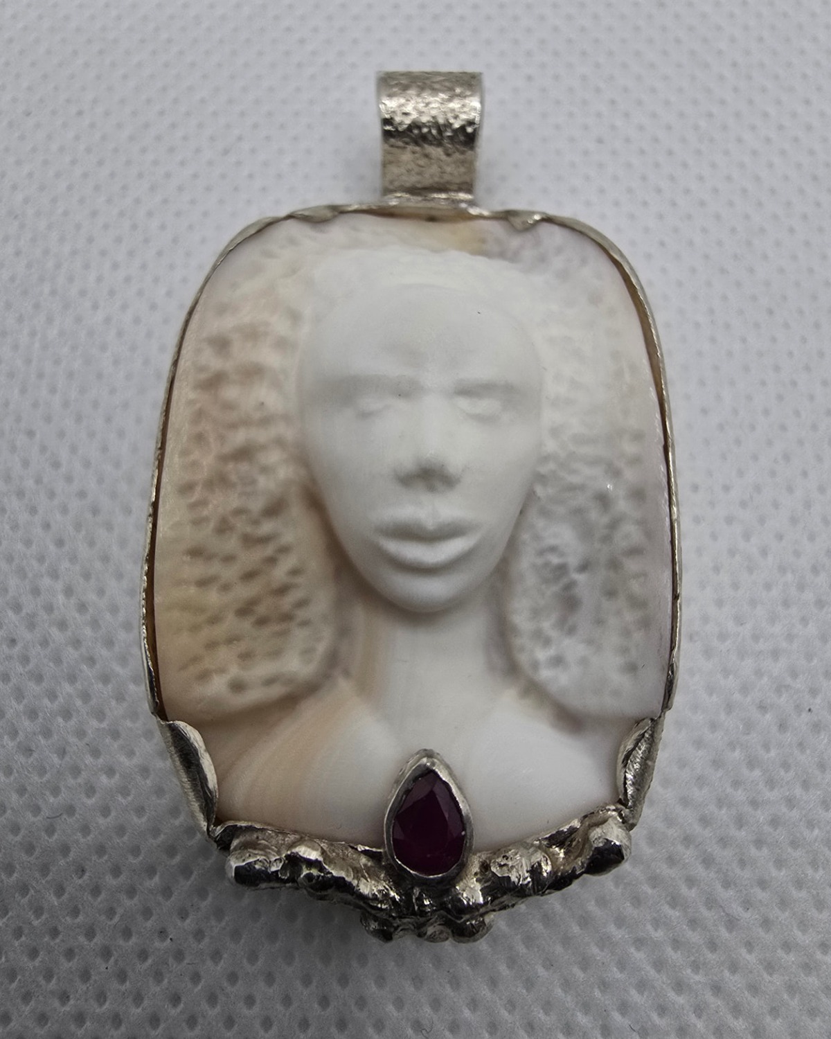 The Ruby Muse — hand-carved natural sea shell cameo of a female figure in frontal high relief, set in handcrafted silver with a pear-shaped faceted ruby beneath the figure