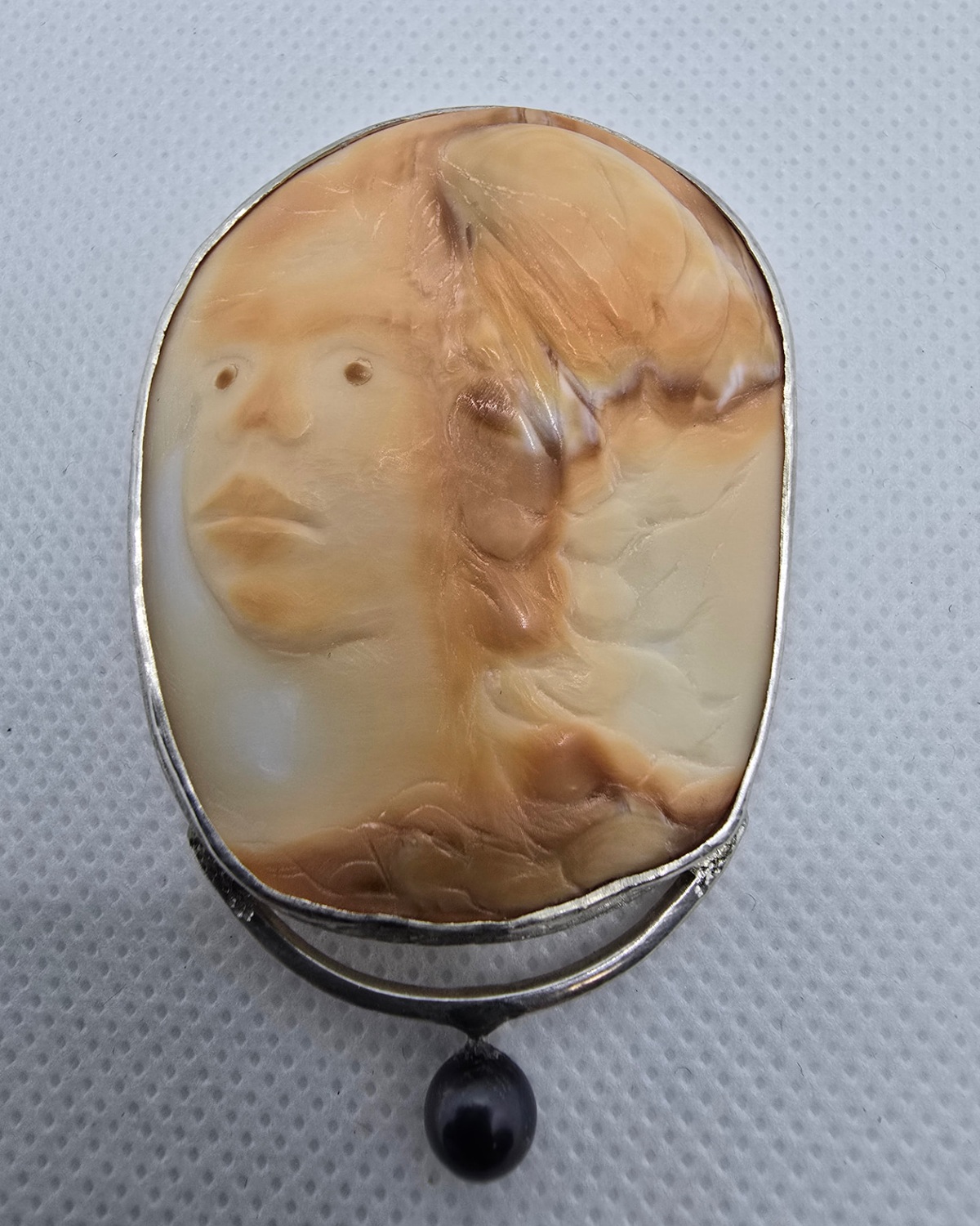 The Guardian — hand-carved shell cameo showing a stylized human face in natural peach and cream shell, with a suspended dark bead below